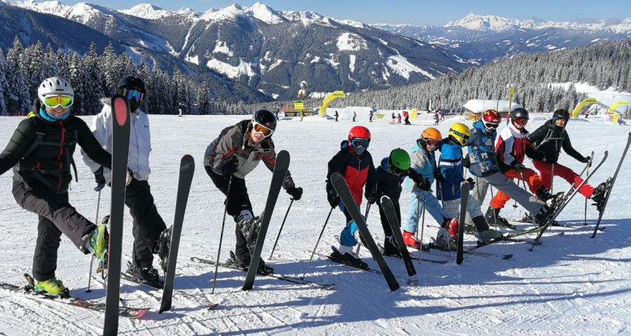 Eine Gruppe Kinder mit ihren Erziehern stehen in Ski-Kleidung in einer Reihe und heben jeweils einen Skier hoch. Sie stehen im Schnee. Im Hintergrund kann man die Berge sehen. Das Bild entstand zu unserer Ski-Freizeit.
