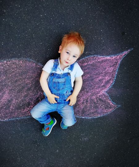 Ein kleiner Junge mit roten Haaren und blauer Jeans-Latzhose liegt auf dem Boden. Auf dem Boden sind mit Kreide Schmetterlingsflügel gemalt. Es sieht aus, als wäre das Kind ein Schmetterling. Der Junge schaut verträumt den Betrachter an.
